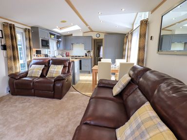 A living room with two brown leather sofas and a kitchen with grey cabinets and a dining table with white chairs at Squirrels Snug in Pony Meadow 1 White Cross Bay near Windermere