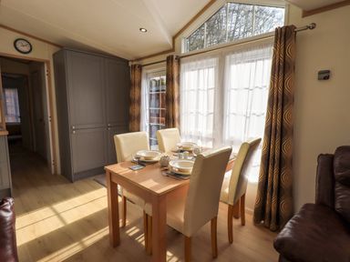A dining area with a wooden table set with dishes and four cream chairs by sheer curtains and a window at Squirrels Snug in Pony Meadow 1 White Cross Bay near Windermere
