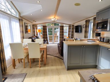 A dining table with four chairs next to a kitchen area and a living room with sofas and a wall-mounted TV at Squirrels Snug in Pony Meadow 1 White Cross Bay near Windermere