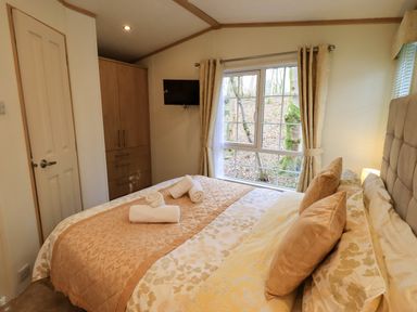 A bedroom with a double bed, pillows, rolled towels, a wardrobe, a window with curtains and a wall-mounted TV at Squirrels Snug in Pony Meadow 1 White Cross Bay near Windermere