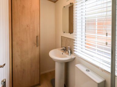 A bathroom with a pedestal sink a mirror a wooden cabinet and a window with blinds at Squirrels Snug in Pony Meadow 1 White Cross Bay near Windermere