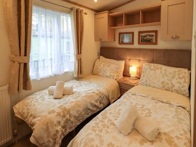 A bedroom with two single beds with floral bedspreads and towels a bedside table with a lamp and wall cabinets at Squirrels Snug in Pony Meadow 1 White Cross Bay near Windermere