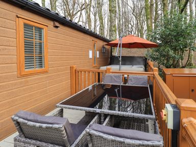 A wooden deck with a glass dining table and chairs next to a hot tub under an orange umbrella at Squirrels Snug in Pony Meadow 1 White Cross Bay near Windermere