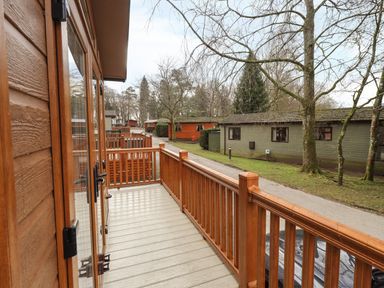 A wooden balcony overlooking a pathway and several cabins with trees around at Squirrels Snug in Pony Meadow 1 White Cross Bay near Windermere