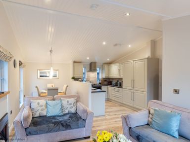 An open living area with two sofas and a dining table next to a kitchen with cream cabinets at Comraich Beckside 40 Limefitt Park near Windermere