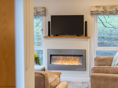 A living room with a wall-mounted TV above an electric fireplace and windows on either side at Comraich Beckside 40 Limefitt Park near Windermere