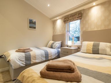 A bedroom with two single beds with striped bedding a window with a floral blind and a nightstand with a lamp and vase at Comraich Beckside 40 Limefitt Park near Windermere