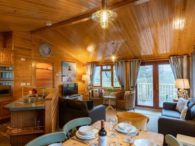 A living room with wood paneling a dining table set with plates glasses and wine a kitchen area sofas armchairs and large windows at Lakeshaven Skiptory Howe 16 White Cross Bay near Windermere