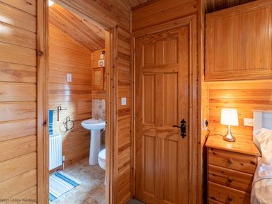 A wooden room showing a doorway to a bathroom with a sink and radiator and a bedside table with a lamp at Lakeshaven Skiptory Howe 16 White Cross Bay near Windermere