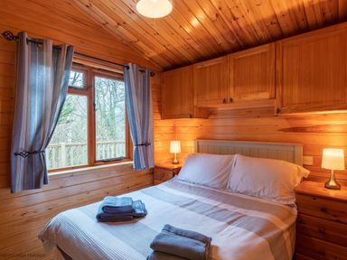 A bedroom with wooden walls ceiling and cabinets a double bed with folded towels two bedside tables and lamps a window with blue curtains at Lakeshaven Skiptory Howe 16 White Cross Bay near Windermere