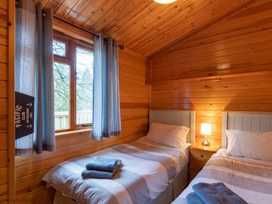 A wooden bedroom with two single beds a bedside table with a lamp and a window with curtains at Lakeshaven Skiptory Howe 16 White Cross Bay near Windermere