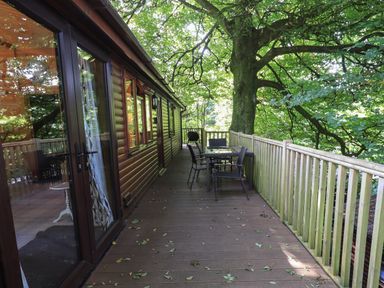 An outdoor wooden deck with a table and chairs next to a cabin with large tree overhead at Lakeshaven Skiptory Howe 16 White Cross Bay near Windermere