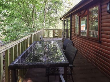 A wooden deck with a glass table and woven chairs outside a wooden cabin at Lakeshaven Skiptory Howe 16 White Cross Bay near Windermere