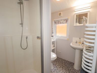 A bathroom with a shower, toilet, pedestal sink, window with a floral blind, small white cabinet, and a towel radiator at Limefitt Lodge Fellside 8 Limefitt Park near Windermere