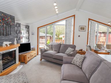A living room with gray sofas a fireplace a wooden TV stand and large windows at Limefitt Lodge Fellside 8 Limefitt Park near Windermere