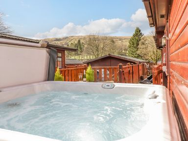 An outdoor hot tub with bubbling water on a wooden deck with a view of wooden cabins and hills at Limefitt Lodge Fellside 8 Limefitt Park near Windermere