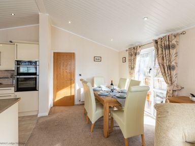 A dining room with a wooden table set for six and cream chairs near a door and window with curtains at Waterbird Lakeview 4 White Cross Bay near Windermere