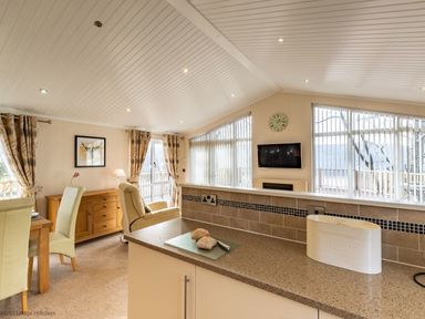 A living area with dining table and chairs a wooden cabinet armchair wall clock television and bread bin on countertop at Waterbird Lakeview 4 White Cross Bay near Windermere