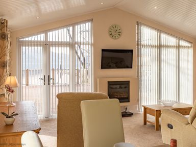 A living room with beige armchairs a wooden table a wall clock a mounted television and large windows with vertical blinds at Waterbird in Lakeview 4 White Cross Bay near Windermere