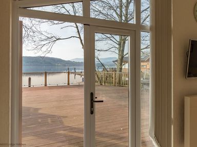 A view through glass doors to a wooden deck with a lake and hills beyond at Waterbird Lakeview 4 White Cross Bay near Windermere