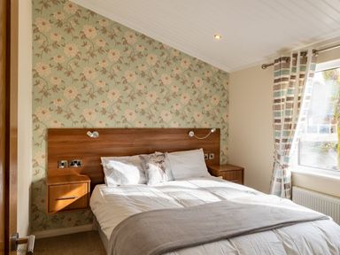 A bedroom with a bed with white bedding and a wooden headboard with attached nightstands and floral wallpaper at Waterbird Lakeview 4 White Cross Bay near Windermere