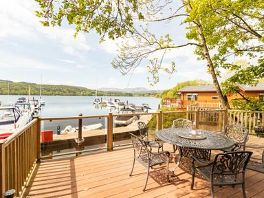A wooden deck with metal table and chairs overlooking boats on a lake with trees and cabins near Waterbird Lakeview 4 White Cross Bay near Windermere