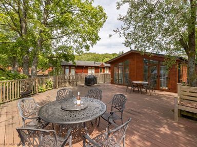 An outdoor wooden deck with metal tables and chairs near wooden lodges and trees at Waterbird Lakeview 4 White Cross Bay near Windermere