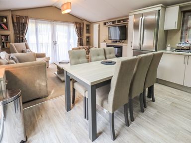 A dining area with six chairs and a table next to a kitchen with a refrigerator and a living room with sofas and a TV at Glenridding Lodge Grasmere 2 White Cross Bay near Windermere