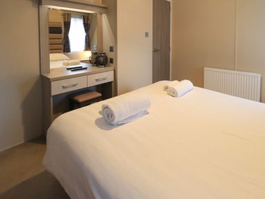 A bedroom with a bed, rolled towels, a dressing table with a mirror and stool, a door, and a radiator at Glenridding Lodge Grasmere 2 White Cross Bay near Windermere