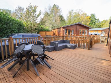 An outdoor wooden deck with stacked plastic chairs around a table and a wicker sofa near wooden cabins at Glenridding Lodge Grasmere 2 White Cross Bay near Windermere