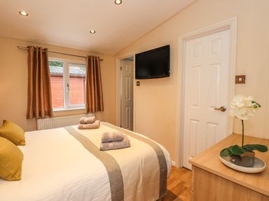 A bedroom with a bed with towels on it a window with curtains a wall mounted television a door and a wooden dresser with a potted flower at Wainwright Lodge Kirkstone 27 Limefitt Park near Windermere