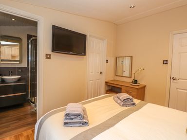 A bedroom with a bed with folded towels a wall-mounted television a wooden desk with a mirror and a potted plant and an open door leading to a bathroom with a sink and shower at Wainwright Lodge Kirkstone 27 Limefitt Park near Windermere