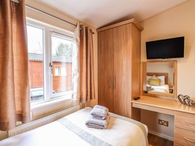 A bedroom with a bed with folded towels a wardrobe a desk with a mirror and a wall-mounted TV at Wainwright Lodge Kirkstone 27 Limefitt Park near Windermere