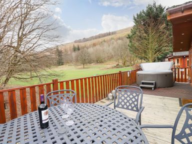 An outdoor deck with a round metal table, chairs, a bottle of wine, two glasses, a hot tub, wooden railing, and a view of trees and hills at Wainwright Lodge Kirkstone 27 Limefitt Park near Windermere