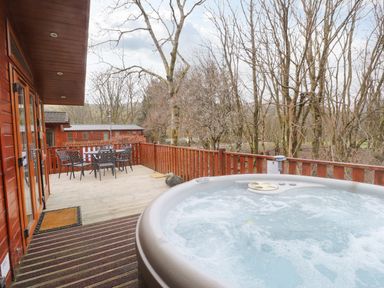 An outdoor wooden deck with a hot tub and a table with chairs surrounded by trees at Wainwright Lodge Kirkstone 27 Limefitt Park near Windermere