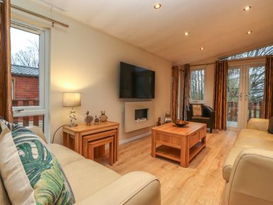 A living room with two beige sofas wooden coffee tables a wall-mounted TV and a window with a view at Wainwright Lodge Kirkstone 27 Limefitt Park near Windermere