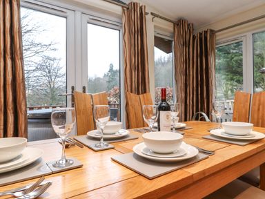 A dining table set with plates bowls wine glasses and a bottle of wine in a room with large windows and curtains at Wainwright Lodge Kirkstone 27 Limefitt Park near Windermere