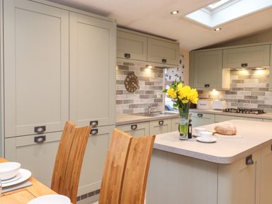 A kitchen with green cabinets wooden chairs a clock on the wall and yellow flowers in a glass vase at Wainwright Lodge Kirkstone 27 Limefitt Park near Windermere