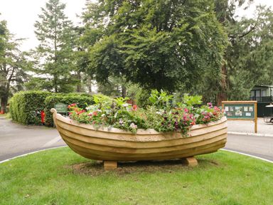 A wooden boat-shaped planter filled with flowers on grass near trees and a road at Beech Grove Lodge in Glade 17 Fallbarrow Park near Bowness on Windermere