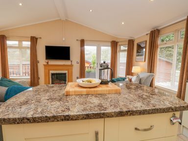 A kitchen counter with a cutting board plate and French press in a living room with sofas a fireplace and windows with curtains at Pheasant's Nest Kirkstone 11 Limefitt Park near Windermere