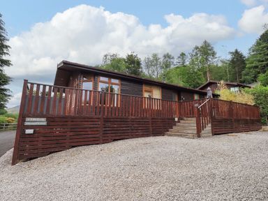 A wooden cabin with a fenced porch and stairs on a gravel area surrounded by trees at Pheasant's Nest Kirkstone 11 Limefitt Park near Windermere