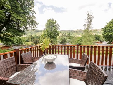 An outdoor wooden deck with a glass top table and wicker chairs overlooking trees and hills at Ambleside Lodge Fellside 3 Limefitt Park near Windermere