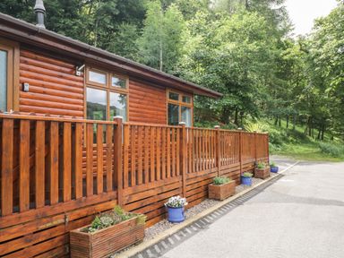 A wooden cabin with a fenced deck and potted plants along a paved path at Ambleside Lodge Fellside 3 Limefitt Park near Windermere