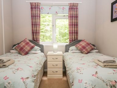 A bedroom with two single beds a nightstand with a lamp and a window with curtains at Ambleside Lodge Fellside 3 Limefitt Park near Windermere