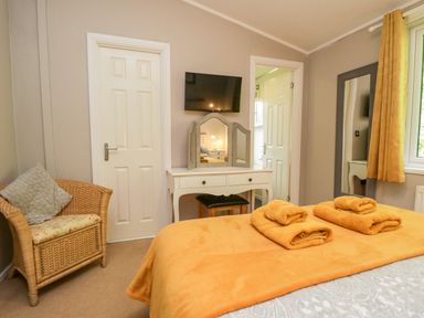 A bedroom with a bed covered in orange blankets and towels a wicker chair a white dressing table with a mirror a wall-mounted TV and a window with orange curtains at Ambleside Lodge in Fellside 3 Limefitt Park near Windermere