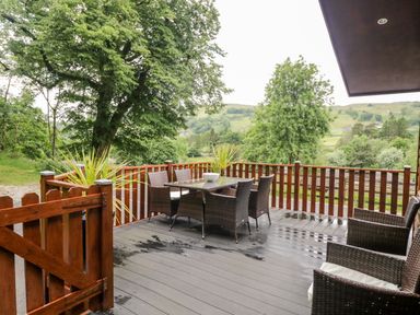 An outdoor deck with a table and six chairs surrounded by wooden railings and trees at Ambleside Lodge in Fellside 3 Limefitt Park near Windermere
