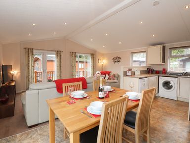 An open kitchen and dining area with a wooden dining table set for four people and a living room with white sofas and red cushions at Robins Nest Bowness 71 White Cross Bay near Windermere