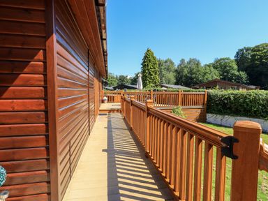 A wooden deck with railing alongside a wooden cabin with trees and neighboring cabins in the background at Robins Nest Bowness 71 White Cross Bay near Windermere