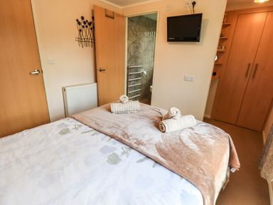 A bedroom with a bed with folded towels on a blanket a wall-mounted TV a radiator and doors at Robins Nest in Bowness 71 White Cross Bay near Windermere