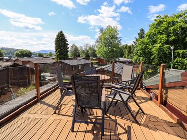 An outdoor wooden deck with six chairs and a round table surrounded by glass panels at Thirlmere View Thirlmere 6 White Cross Bay near Windermere